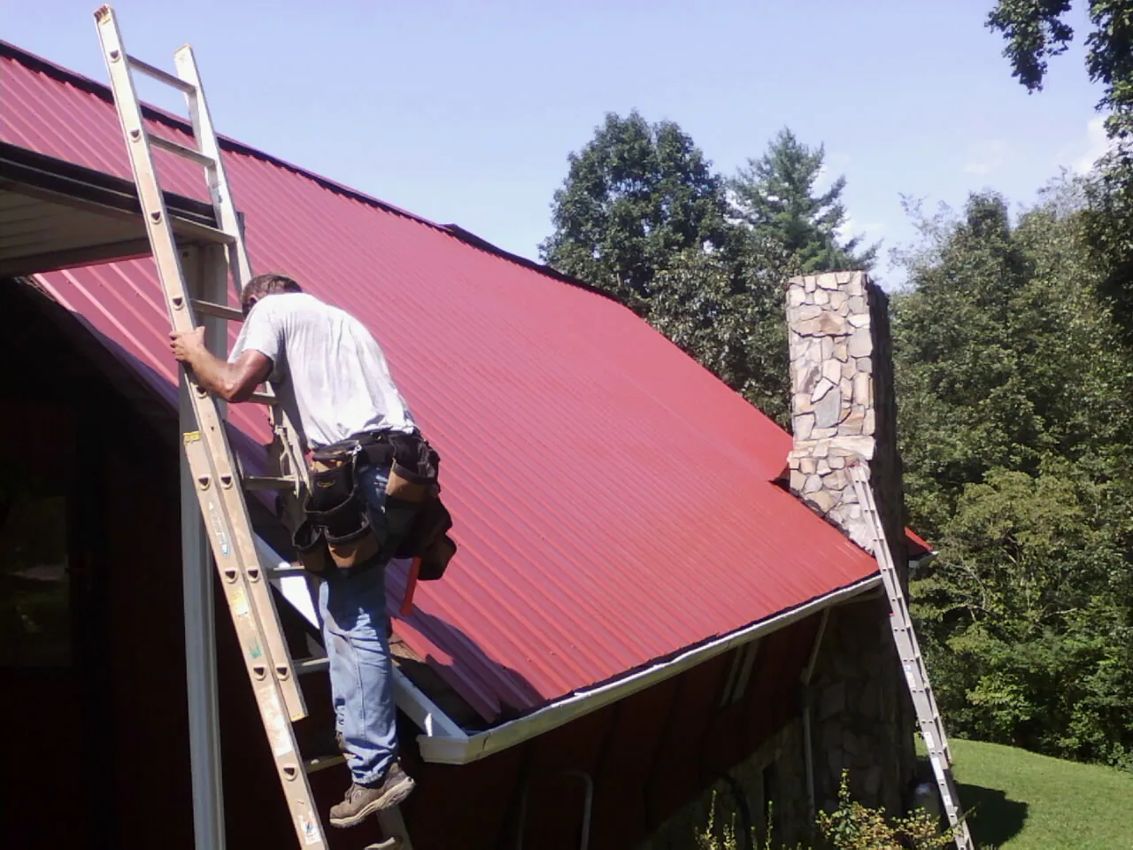 Roofer on a ladder beside a red metal roof and stone chimney — Soffit & Fascia Repair by Roofing Brothers in Cleveland